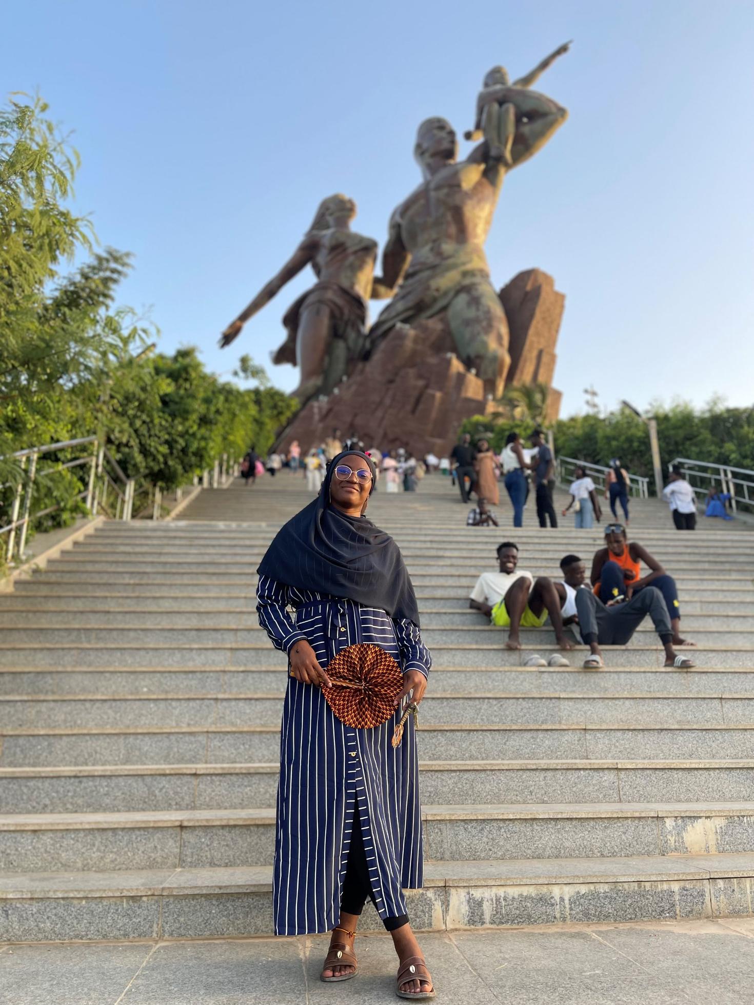 Étudiante africaine posant devants des escaliers montés par des personnes. Deux grandes statues en arrière-plan