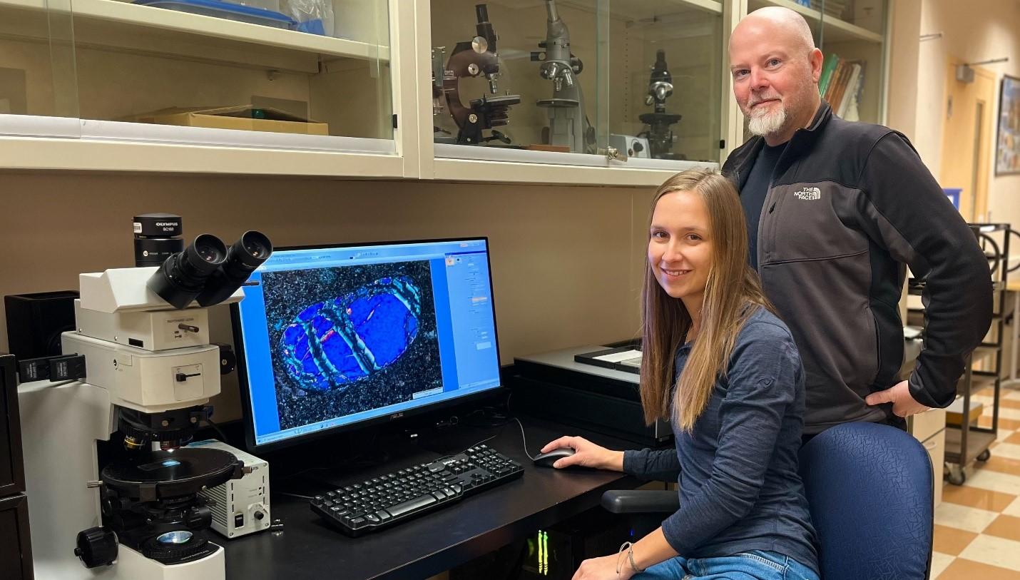 PhD student Jennifer Spalding is sitting at a lab bench in front of a computer and Professor David Schneider is standing to her right. 