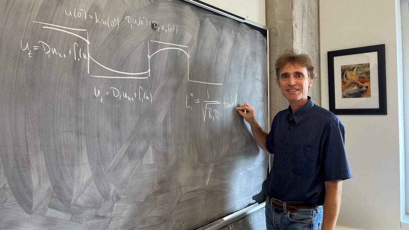 Professor Frithjof Lutscher is standing to the right of a chalkboard on which he is writing a mathematical formula. 