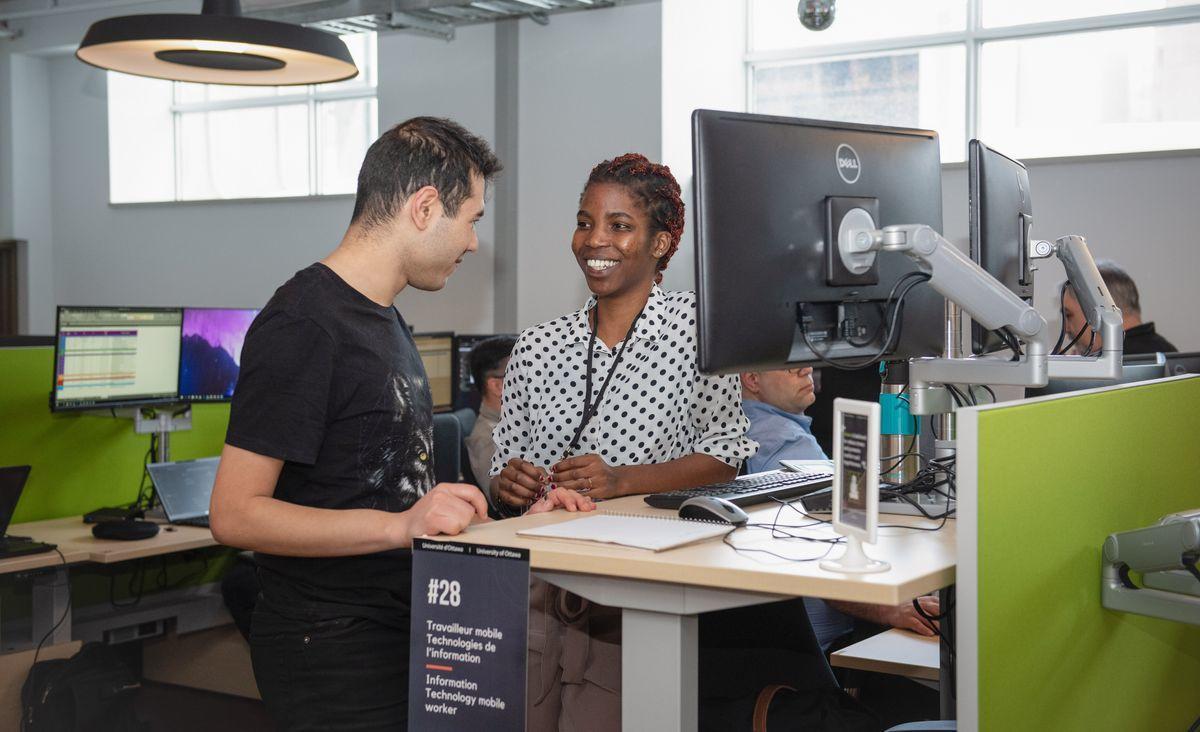 Un homme et une femme sont debouts et discutent devant un bureau de travail surélevé.