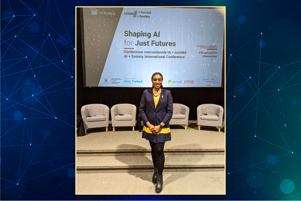 A woman stands in front of a stage. The stage has 4 empty chairs. There is a large screen in the background.