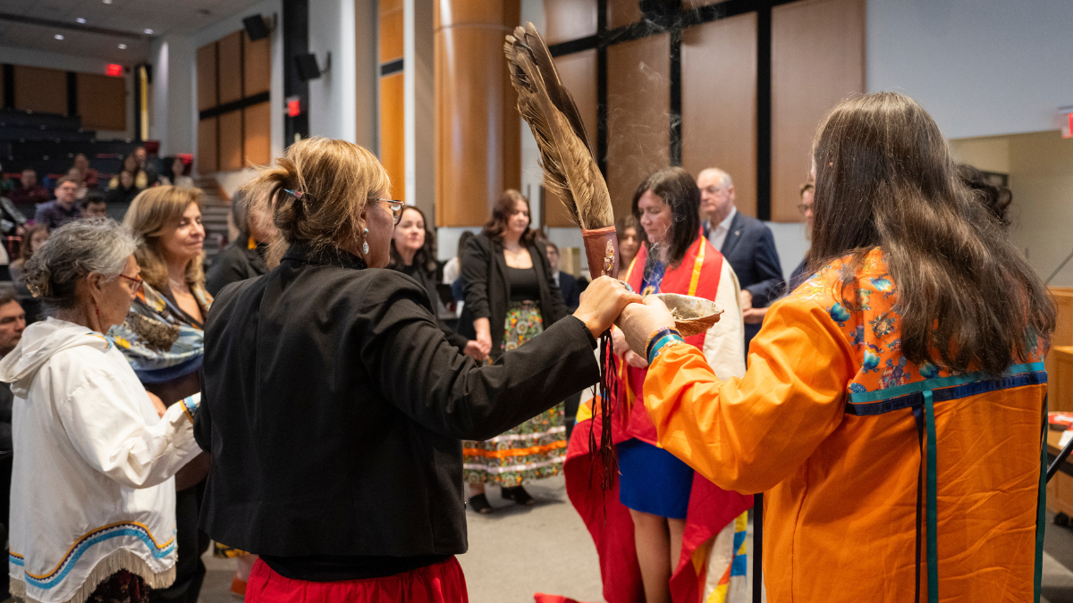 a woman wrapped in a blanket stands in the centre of a group of people who are holding hands. A man in the circle holds an eagle feather
