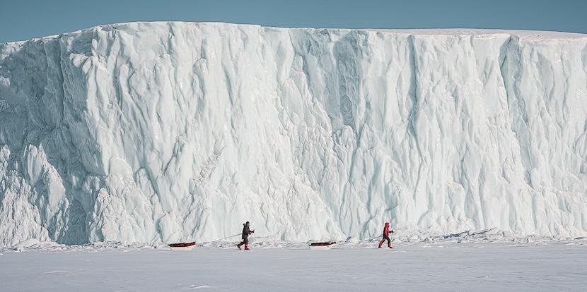 Deux explorateurs tirant leurs traineaux devant une falaise de glace. 