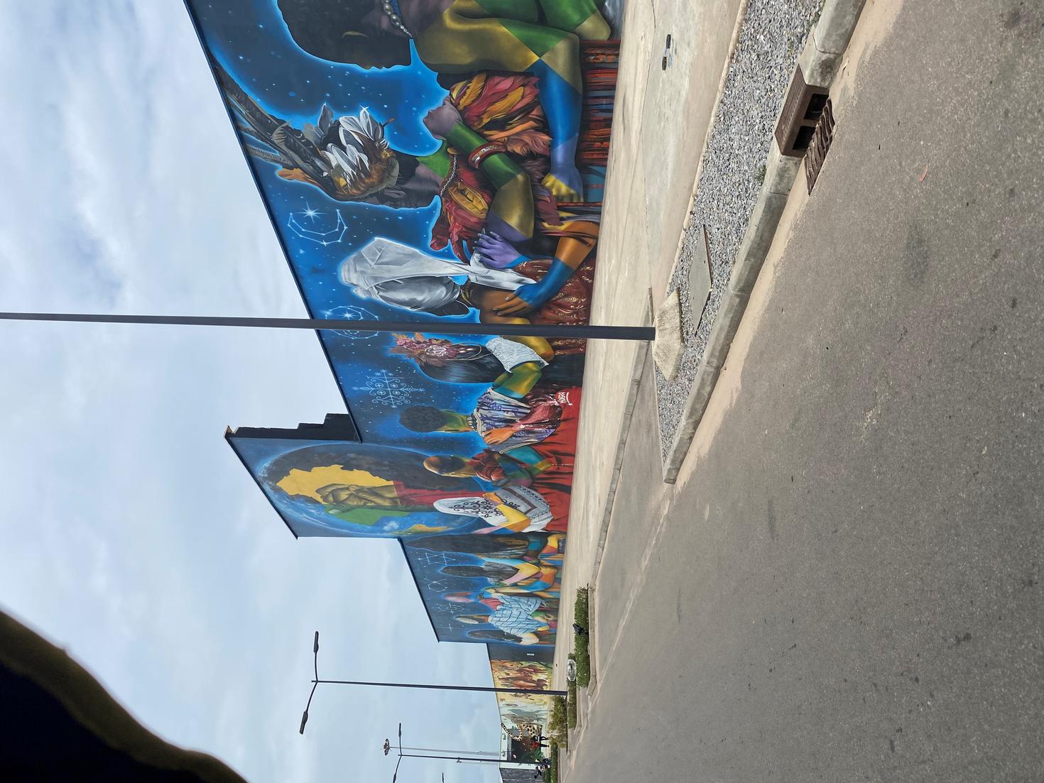 Photo of a sidewalk taken from inside a car. Street lamps in the foreground and a large wall decorated with drawings and graffiti with a blue background in the background.
