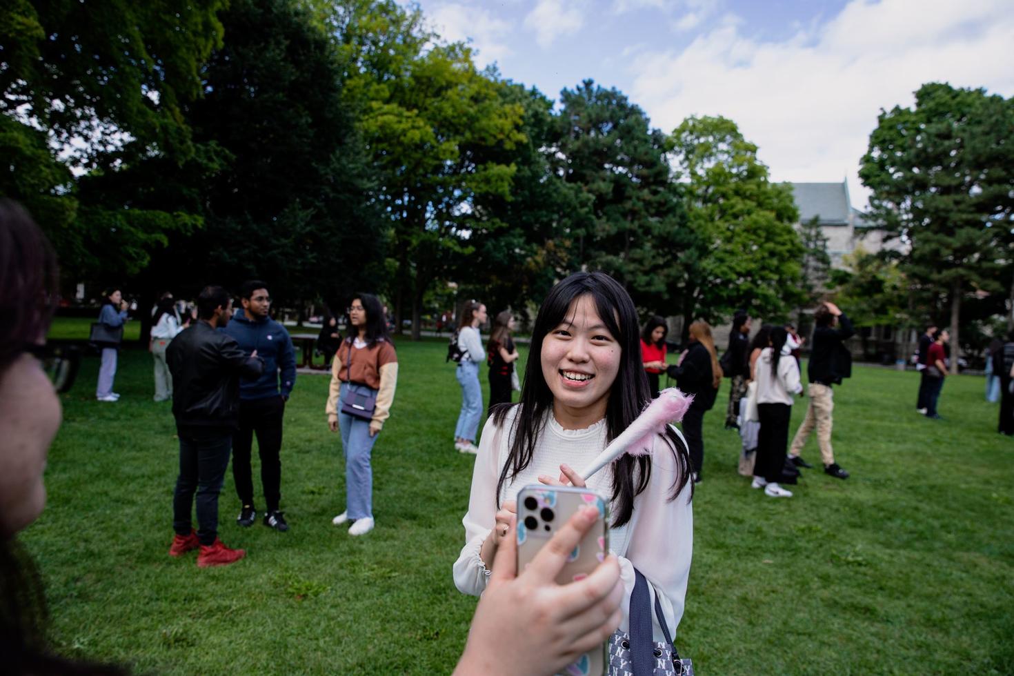 A smiling student stands on the Tabaret lawn while her friend posts a photo with her phone.