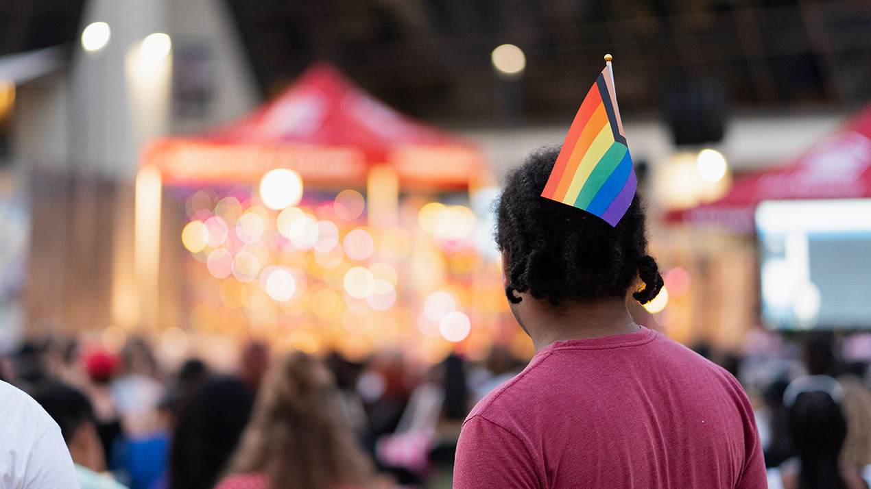 A student with a Pride flag in his hair looks at a brightly lit stage in the distance.