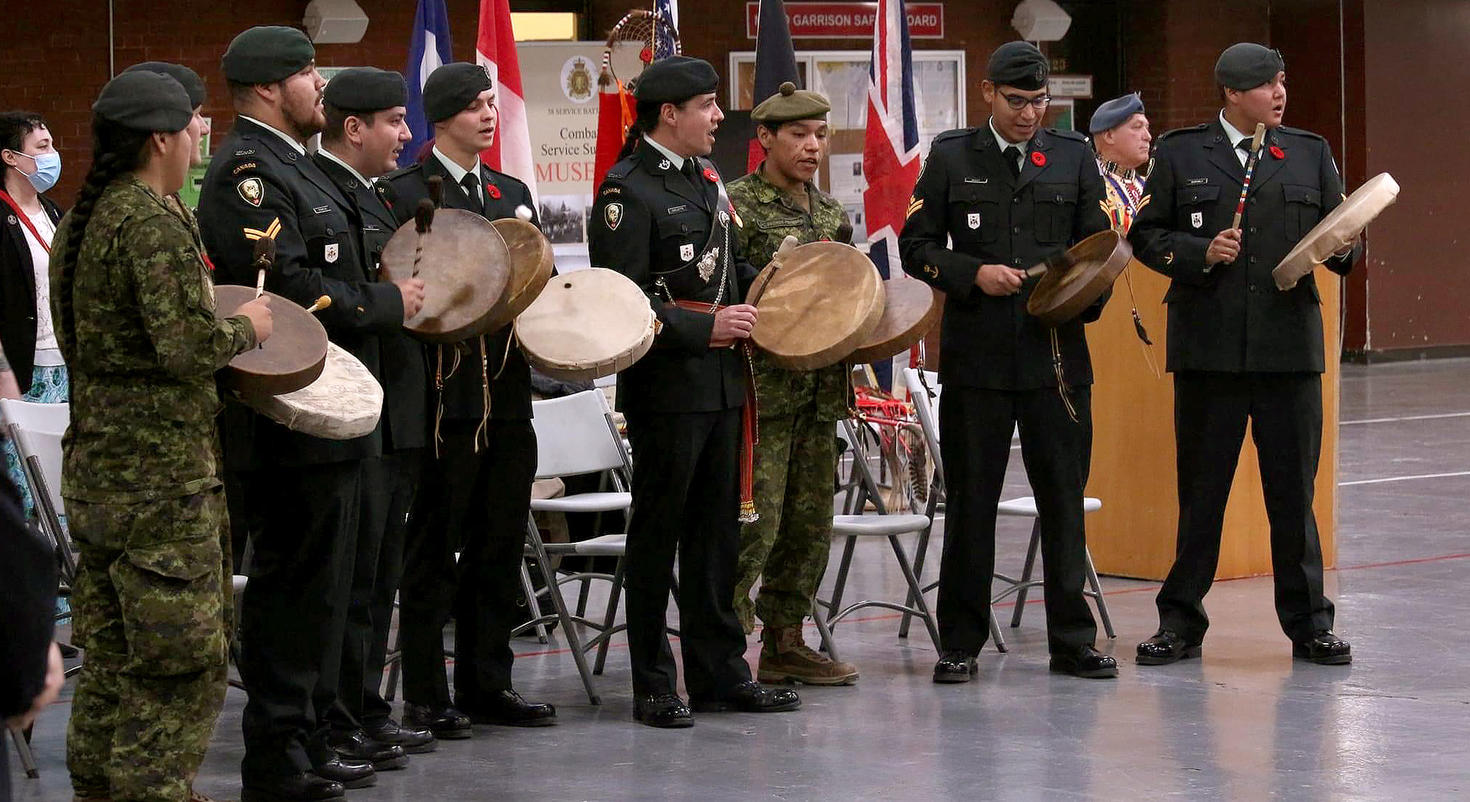 The Warriors of the Royal Winnipeg Rifles (Indigenous drum group) in uniform hold drums and stand together during a ceremony with flags in the background.