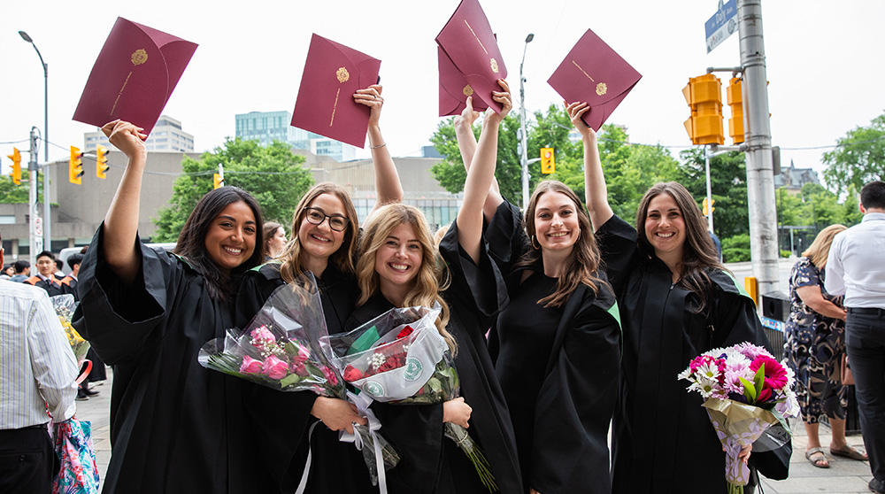 Cinq femmes vêtues de robes de fin d'études brandissent leurs diplômes.