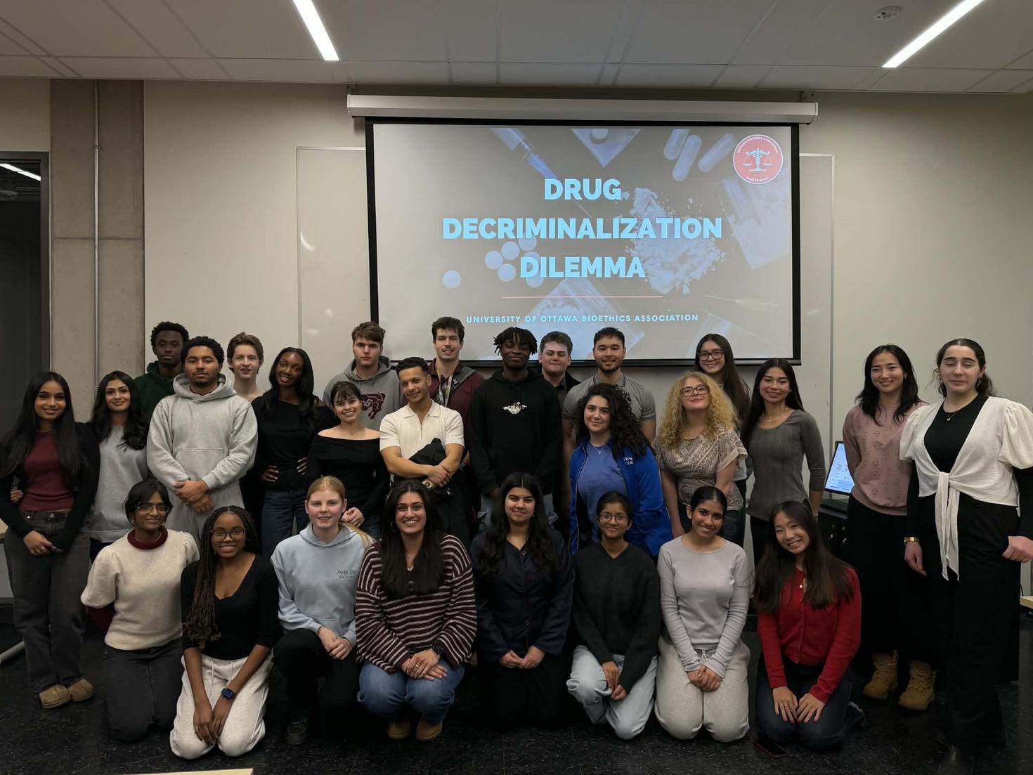 Large group photo of uOttawa Bioethics Association members after their Drug Decriminalization Dilemma event, with a projector behind them displaying the event title and club name and logo over an image of drugs.