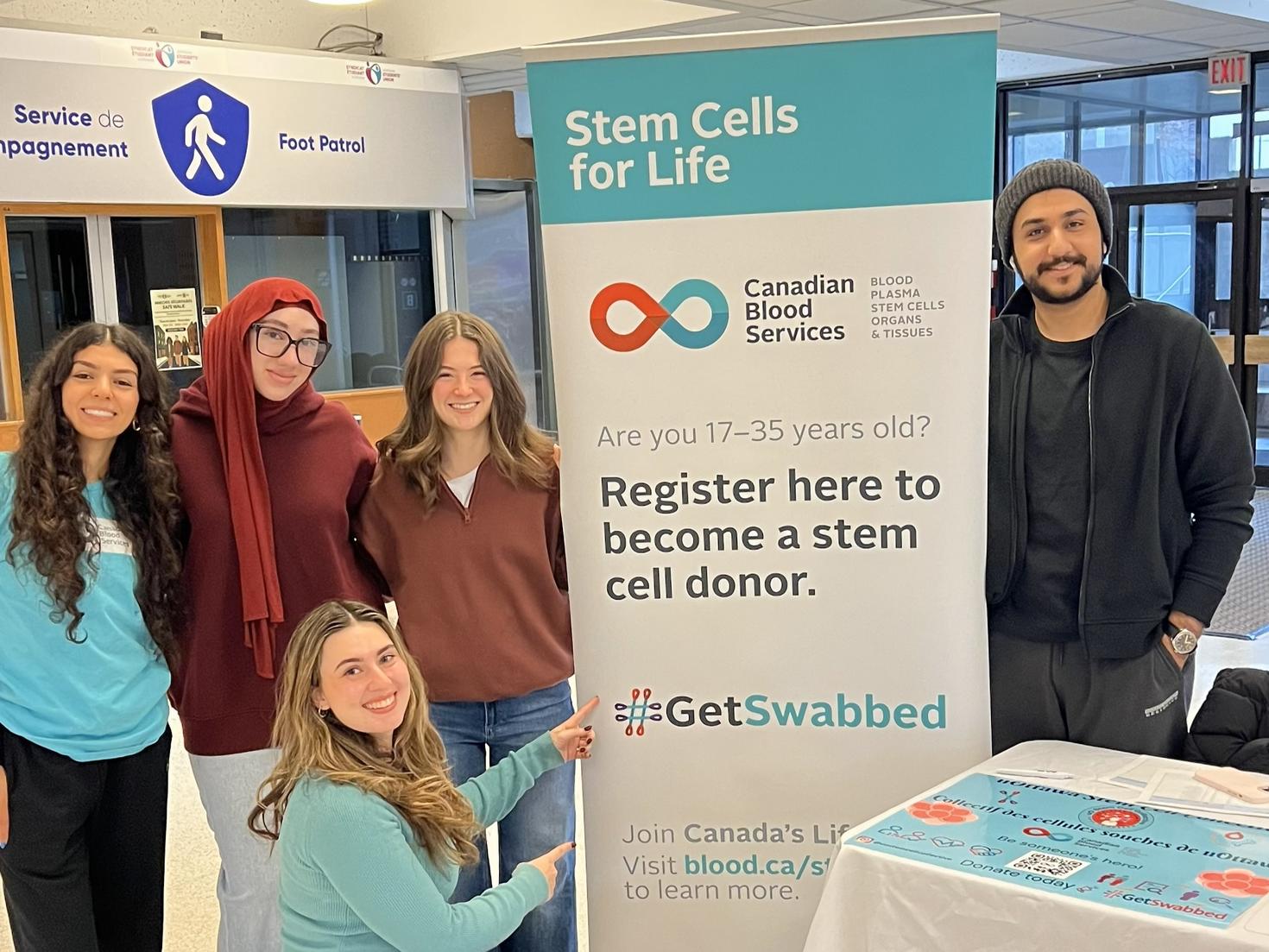 uOttawa Stem Cell Collective executives at the Winter Clubs Fair, posing around a large "Stem Cells for Life" banner from Canadian Blood Services. One executive kneels and points toward the "#GetSwabbed" hashtag, while four others stand alongside the banner and a table featuring the club’s logo and a QR code.