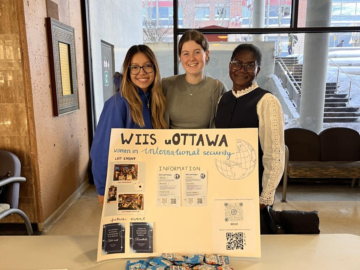 uOttawa Women in International Security executives at the Winter Clubs Fair, standing behind a large white display board on a table covered in packets of Welch's fruit snacks and flyers. The board showcases photos of past and future events, information about the club, and an Instagram QR code.