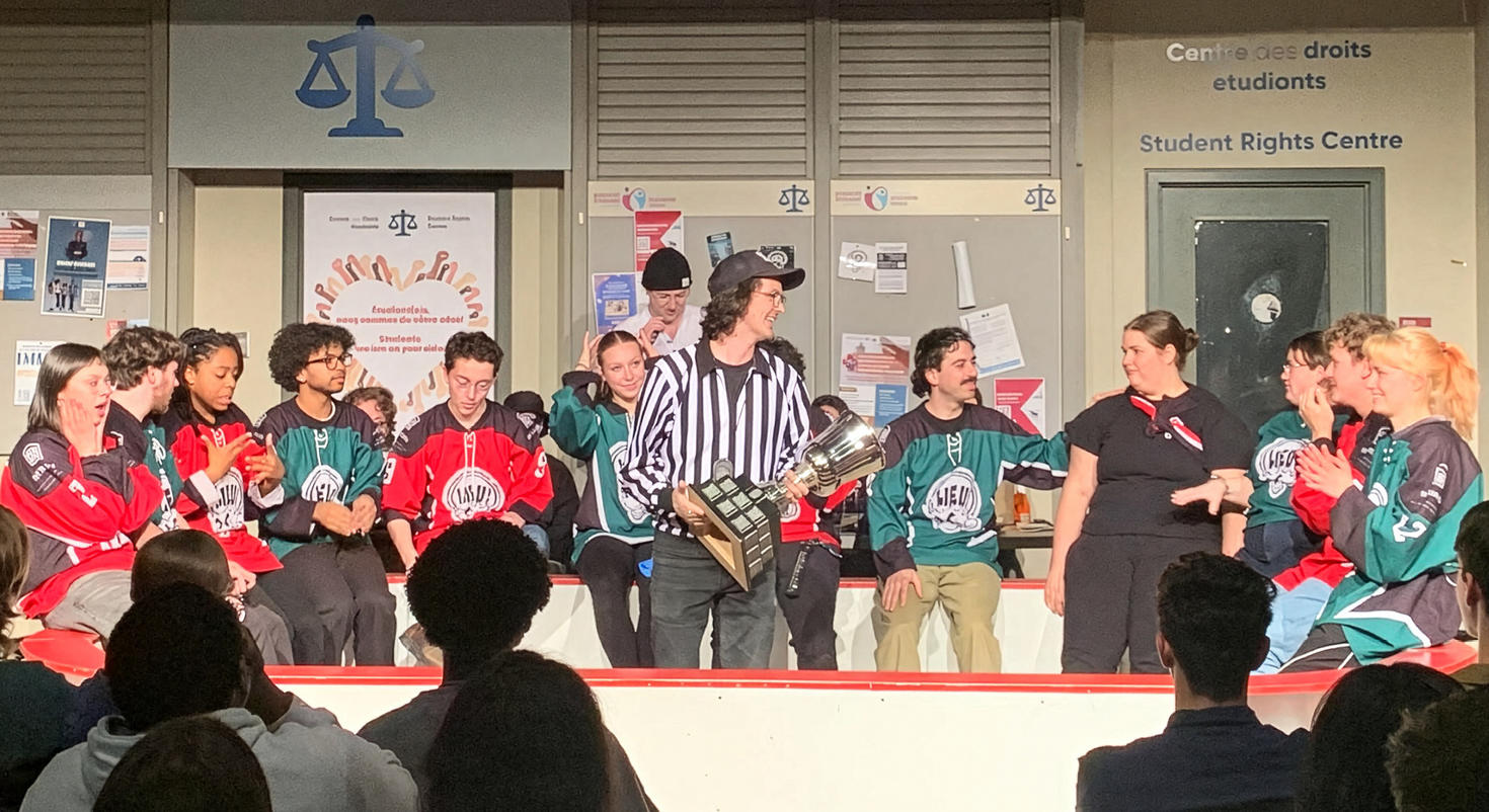 Les membres du club d'improvisation LIEU, vêtus de maillots de hockey, sont assis sur la scène de l'UCU Terminus pendant un match d'improvisation. Au centre, un artiste déguisé en arbitre tient un grand trophée.