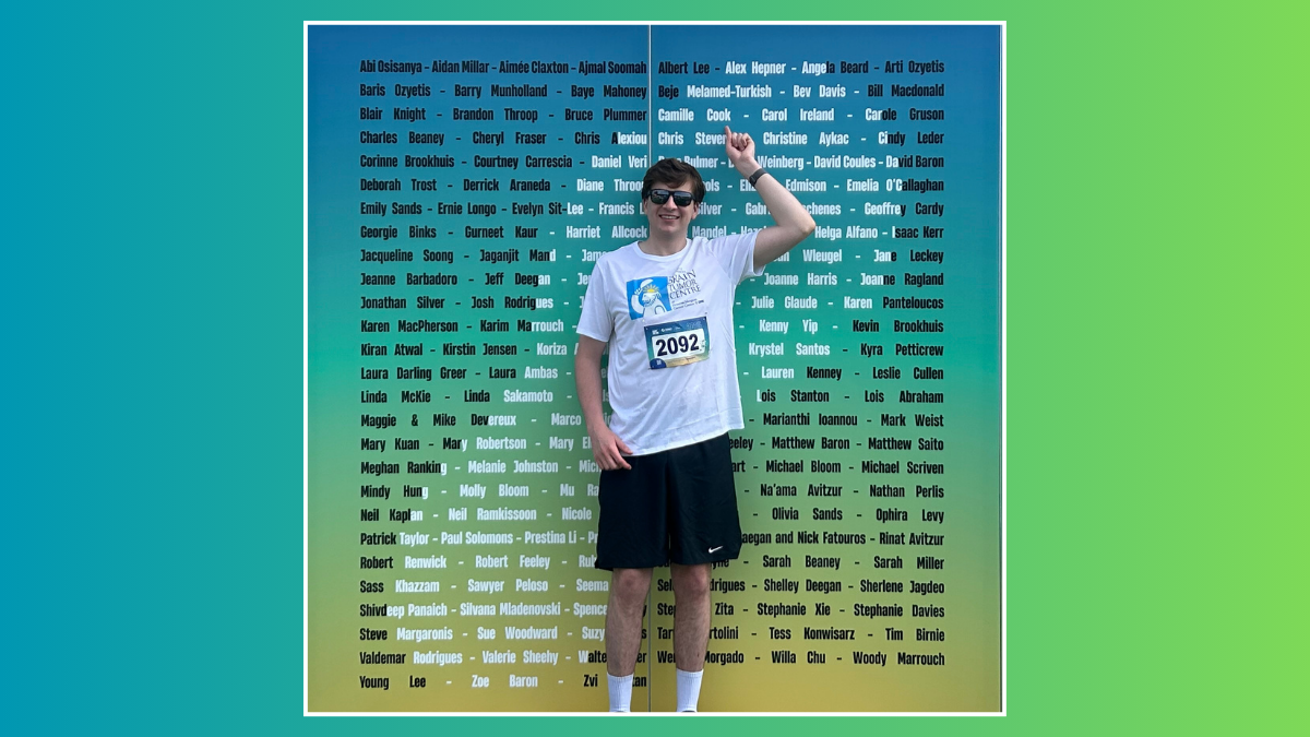 A university aged male, wearing a Tshirt, black shorts and sunglasses, stands against a billboard with a list of names and points. The name he is pointing at is Alex Hepner.