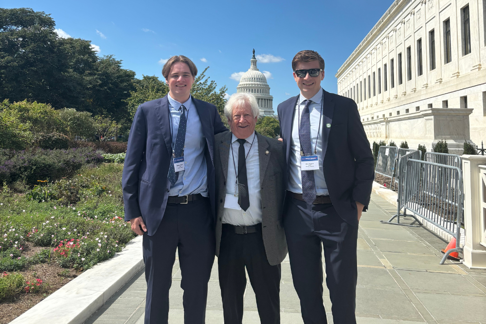 Three men in suits, two younger and one older, stand outside and smile at the camera. In the distant background, the US Capitol building is visible.