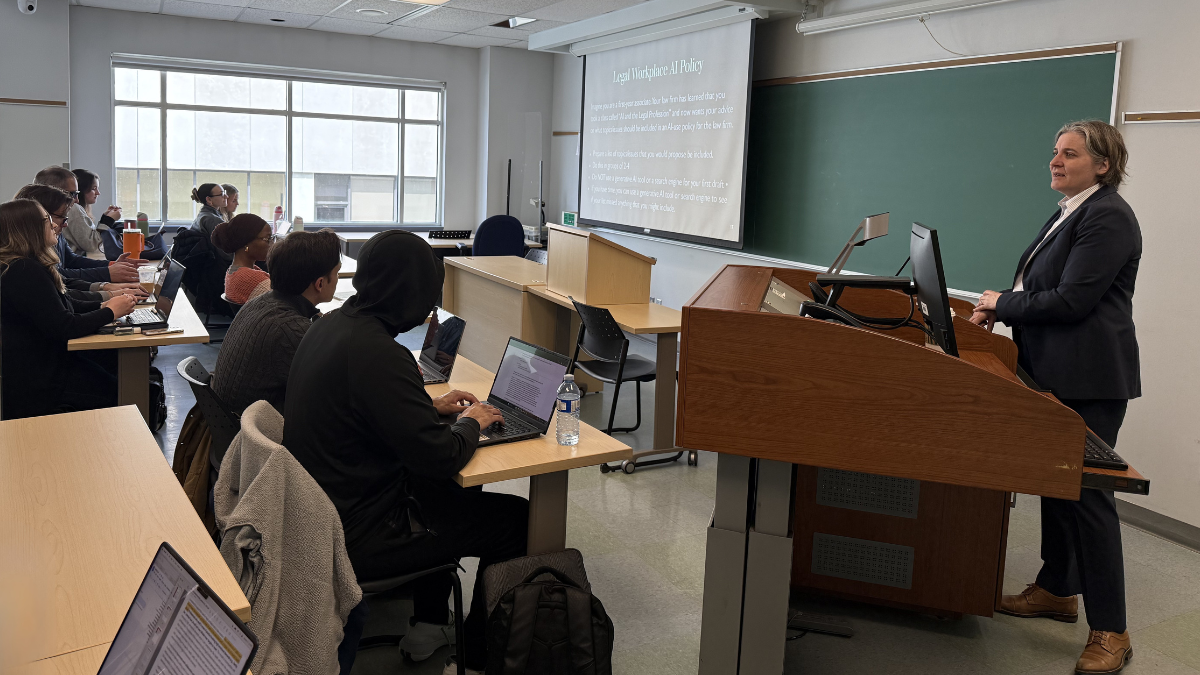 A professor stands at the front of the class, facing students sitting at desks. The students are looking at a large screen at the front of the class.