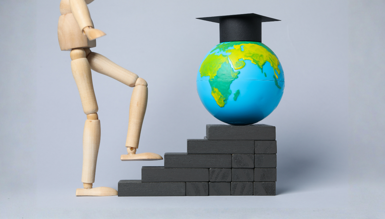 figurine en bois sur le point de monter des marches en direction d'un globe terrestre, coiffée d'un mortier, symbolisant le parcours universitaire