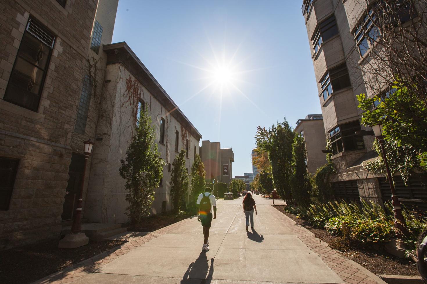 Two students walk towards the shining sun along the Grand Allée | Kichi Mìkàns on the University of Ottawa campus.