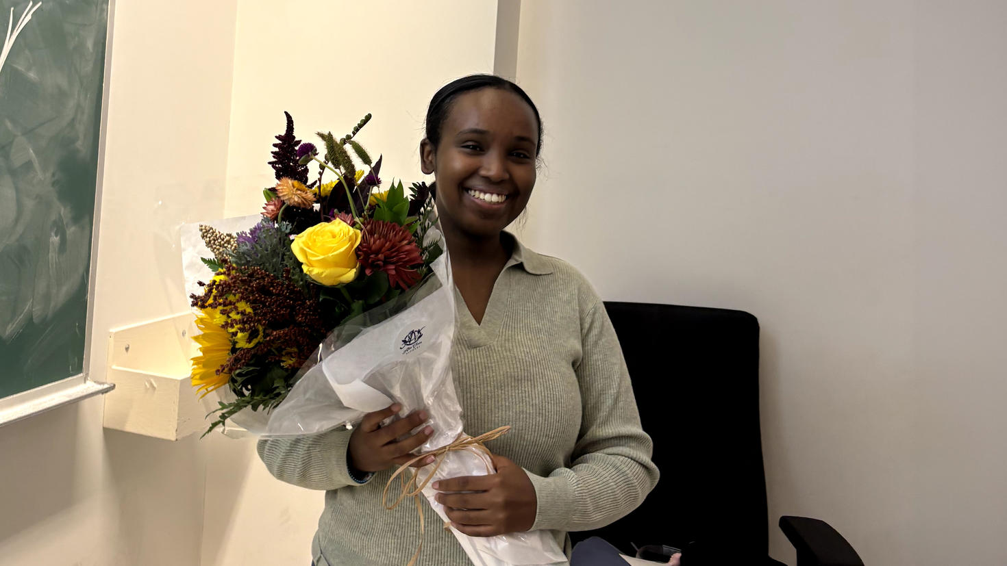 A person standing in front of a chalkboard, smiling and holding a bouquet of flowers.