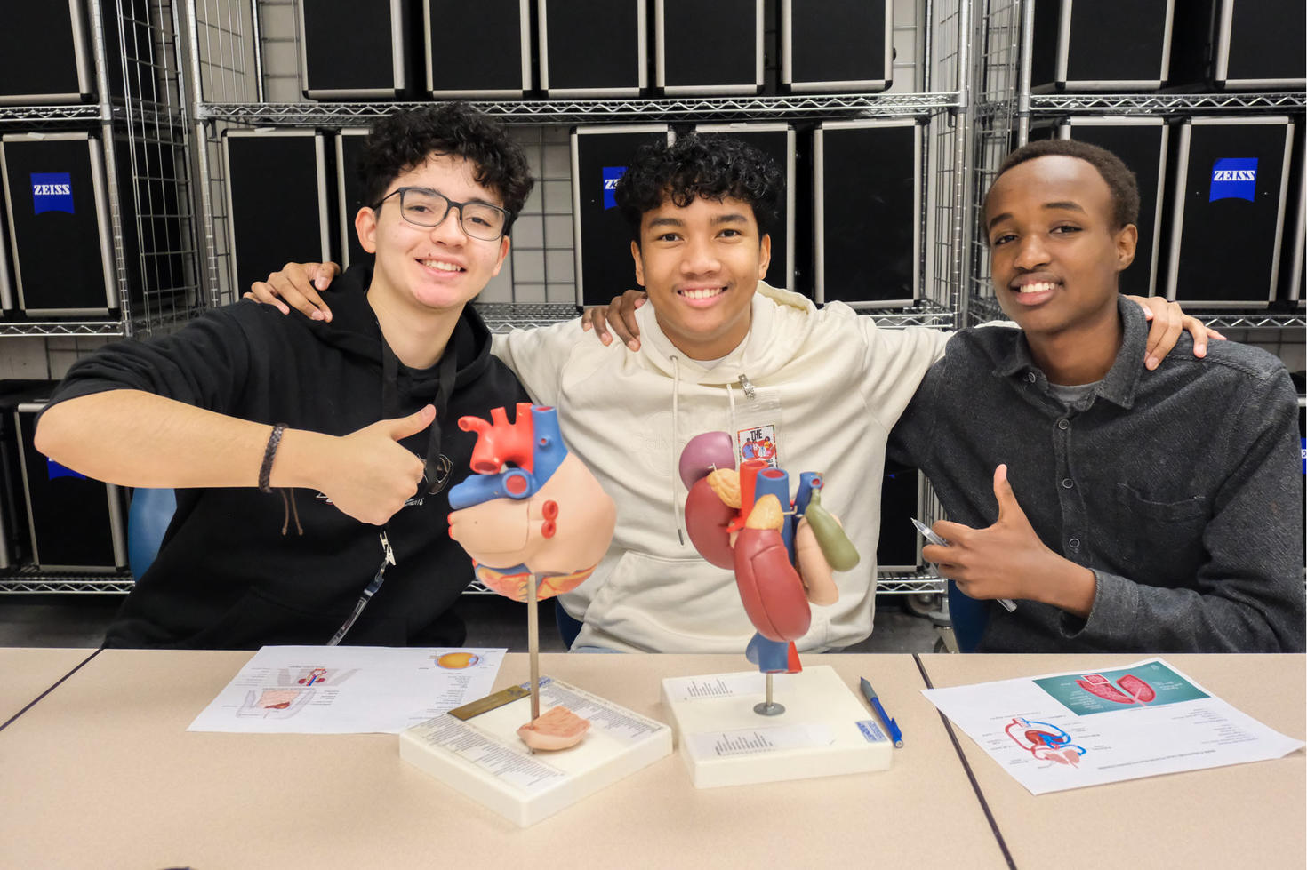Three young students smiling as they work with anatomical models on a desk.