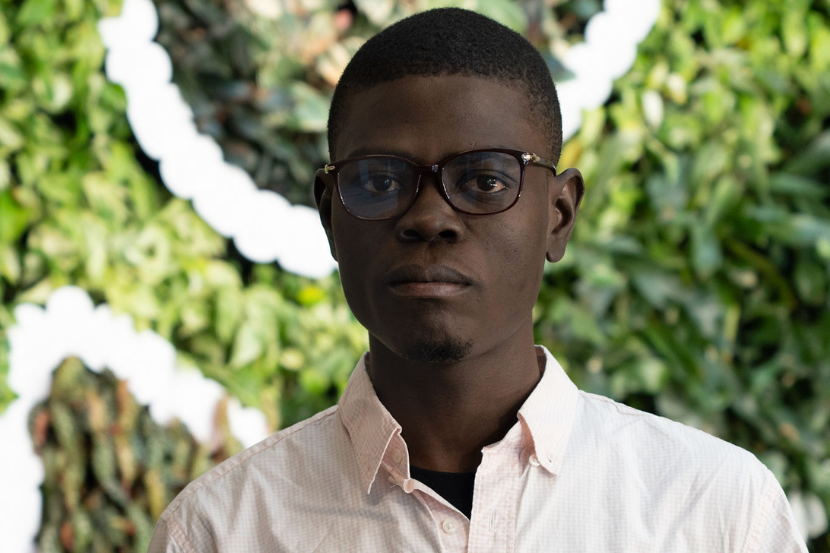 Portrait of Pierre Jameson Beaucejour wearing glasses and a light-coloured button-up shirt, standing in front of a green living wall with white decorative elements. He faces the camera with a neutral expression.