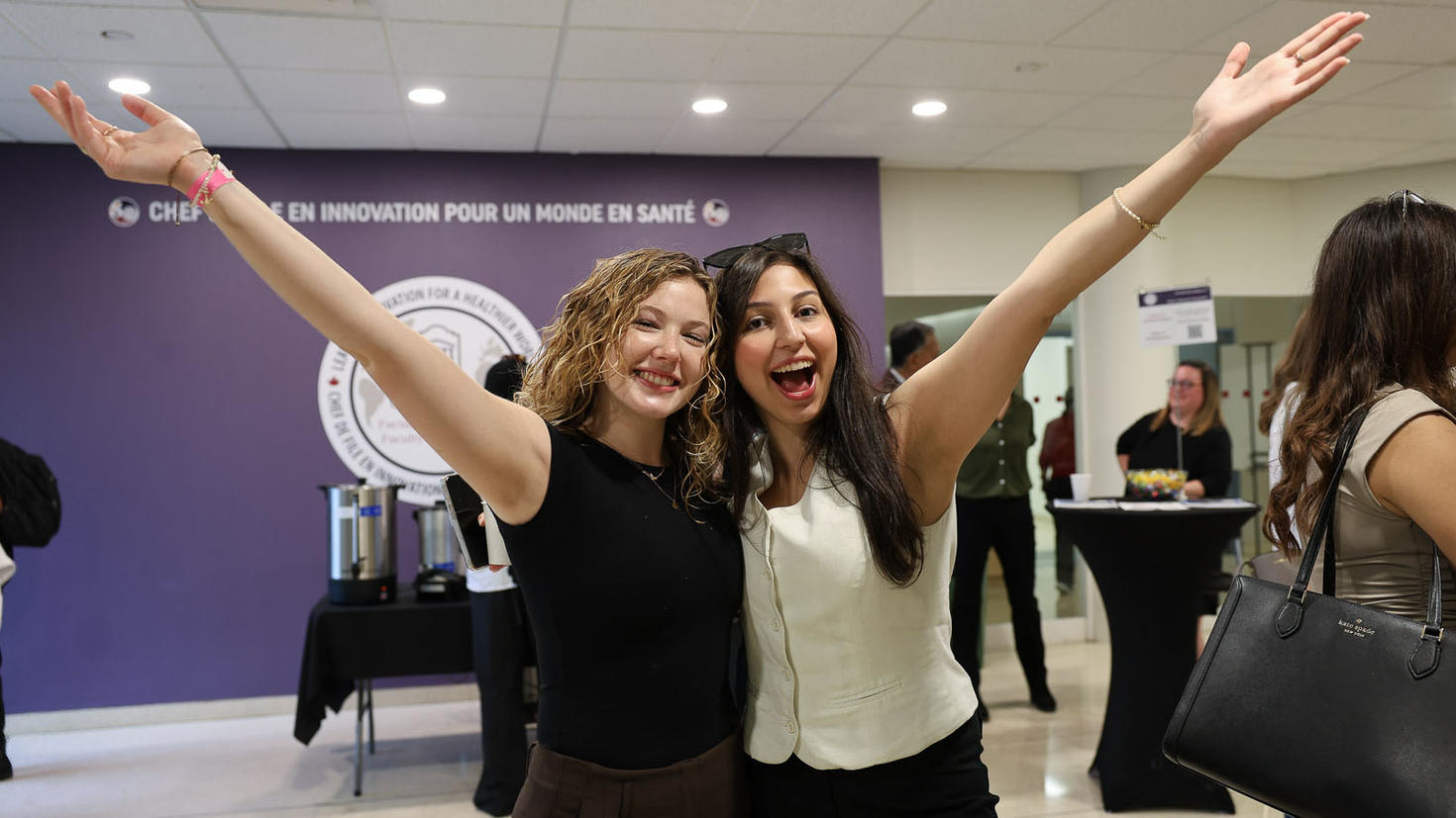 Deux étudiantes sourient, se prennent dans les bras et font signe à la caméra.