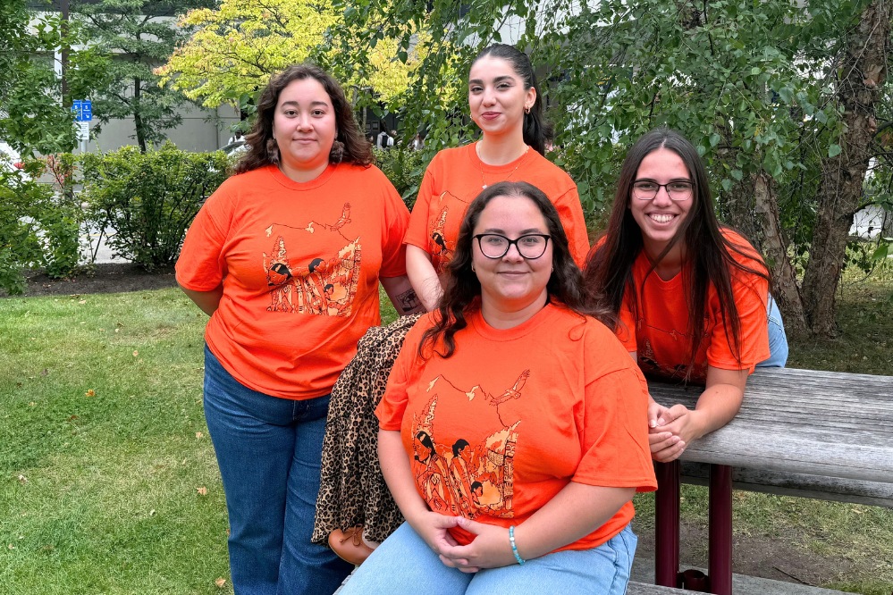 Quatre jeunes femmes, toutes vêtues d’un chandail orange au même motif, sont assises ou debout près d’une table de pique-nique à l’extérieur.