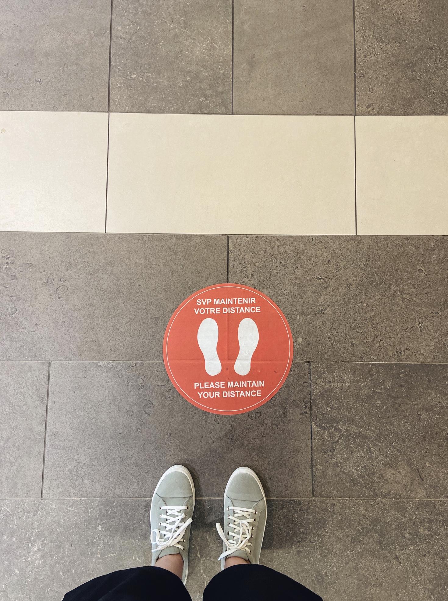 the shoes of a person standing next to a sticker on the floor that indicates to keep one's distance