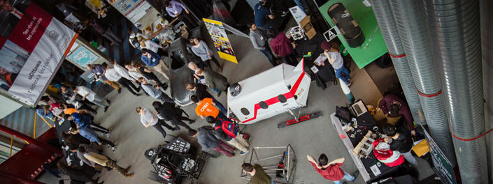 A view from above of around 20 students looking at a covered vehicle with portholes as well as two cars in the SITE lobby.