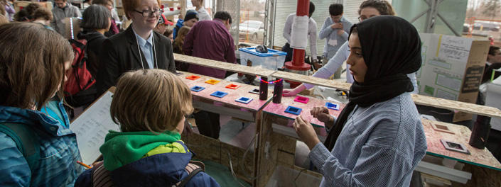 A woman speaking to three students in front of a long table that holds square pots.