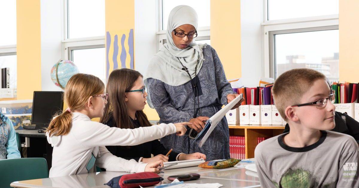 Children sit at their desks in a classroom. Two of them interact with a teacher.