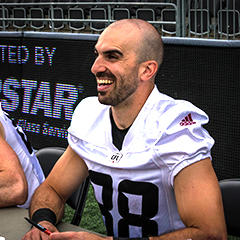 Brad Sinopoli sits at a table smiling and signing autographs for fans.