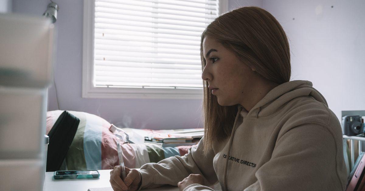 A young woman seen from the side. She's sitting at a desk in her bedroom.