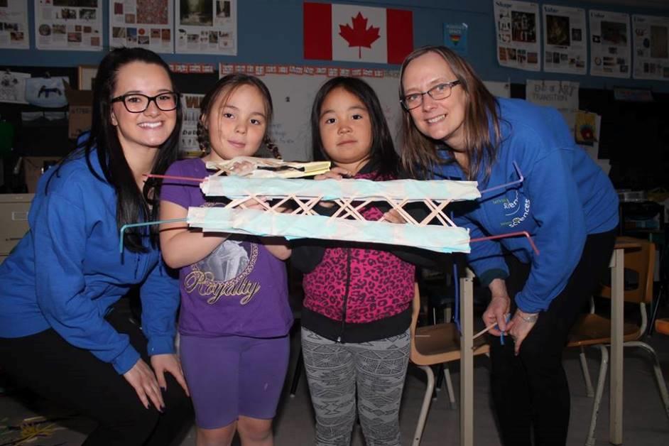 Two women pose with two girls holding a paper model of a bridge.