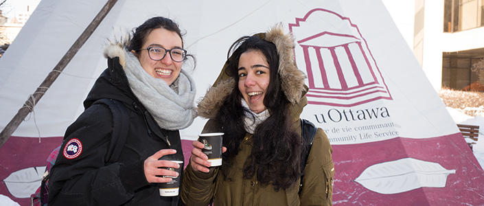 Two students smiling in front of a uOttawa branded tent holding cups of coffee.