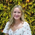 Une femme blanche avec des cheveux blonds et vêtue d'une robe blanche avec des motifs fleuris en face du mur de plantes de FSS