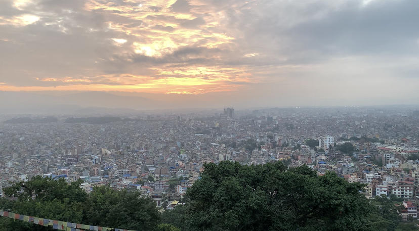 Vue en perspective d'une ville du Népal. Deux arbres sont visibles au premier plan, suivis d'une multitude de bâtiments urbains. Ville couverte par un coucher de soleil