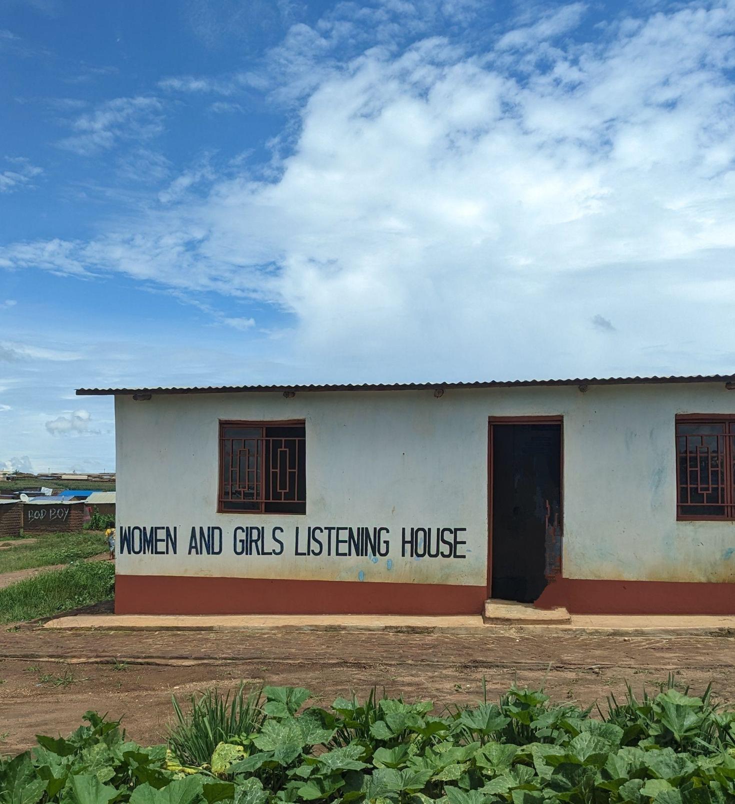 Petit bâtiment au milieu d'un champ. Murs blancs et orange avec l'inscription « Maison d'écoute pour femmes et jeunes filles » sur un mur. Ciel bleu en arrière-plan