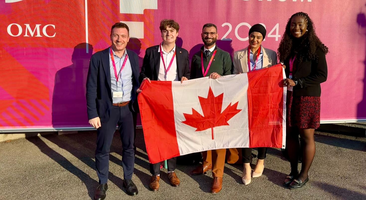 Cinq membres de la délégation de Jeunes diplomates du Canada au Forum public de l’Organisation mondiale du commerce (OMC) à Genève. Joshua Finkelstein (Université de Reading, R.-U.), Émile Cloutier (Université Laval), Elie Kallab (Université d’Ottawa), Sahib Kaur Dhaliwal (diplômée de l’Université d’Ottawa) et Angelica Kalubiaka (Université d’Ottawa) sont devant une bannière de l’OMC arborant un drapeau canadien.
