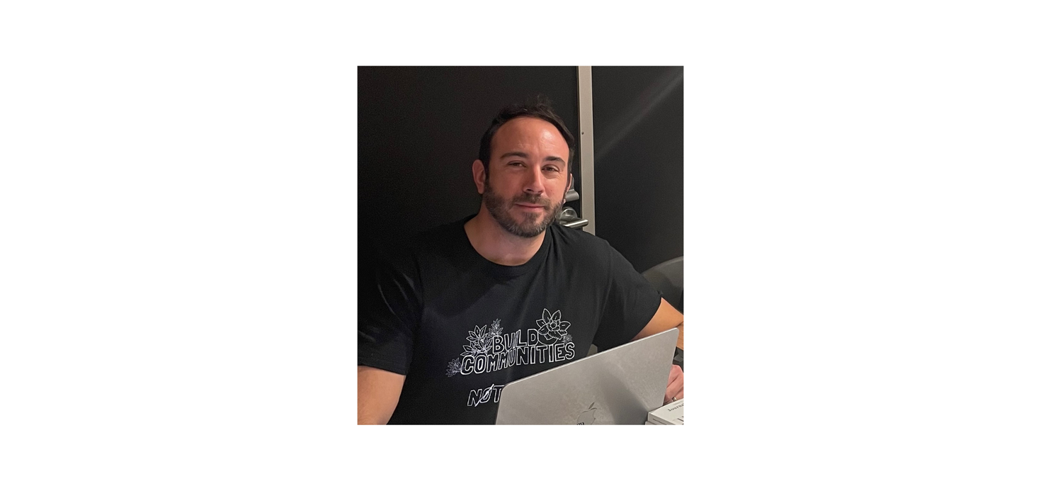 Une personne aux cheveux bruns courts et à la barbe est assise à une table avec un ordinateur portable. Elle porte un t-shirt noir sur lequel est imprimé « Build Communities ».