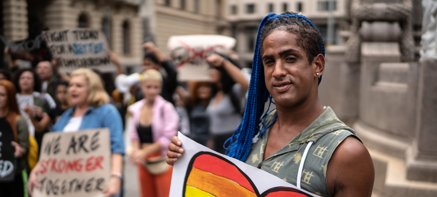 Une personne tenant un cœur arc-en-ciel devant une foule de manifestants. Une femme tient une pancarte sur laquelle on peut lire : « Ensemble, nous sommes plus forts ».