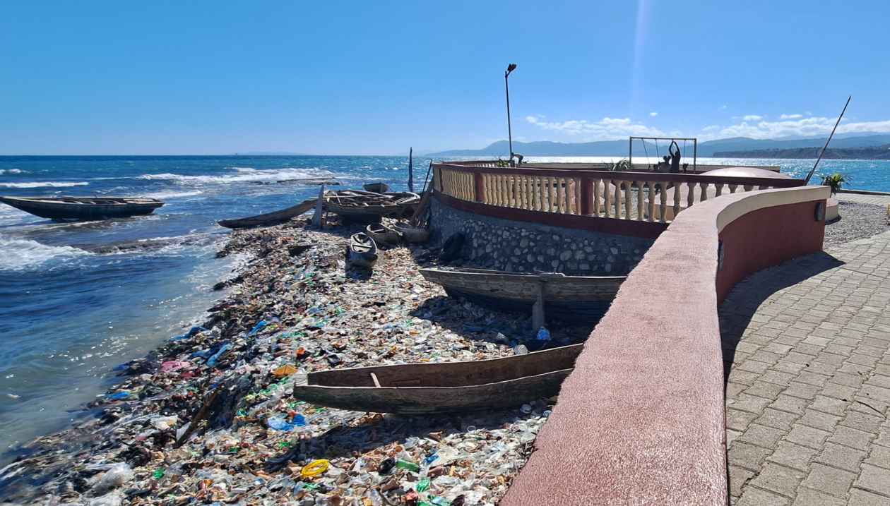 Vue du littoral du Fort La Pointe à Jérémie, en Haïti. Plusieurs embarcations de pêche en bois reposent sur le rivage, à côté d'une importante accumulation de déchets plastiques et d'autres détritus. Une promenade rénovée bordée de murets en pierre surplombe la plage, illustrant le contraste entre la mise en valeur du patrimoine, la pêche artisanale et les défis environnementaux persistants.
