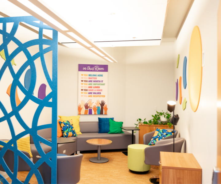 Un espace de détente accueillant comprenant des fauteuils modernes, des coussins colorés, des plantes et des éléments décoratifs muraux. Une grande affiche au fond de la pièce présente des messages valorisants, notamment « Vous avez votre place ici », « Vous comptez » et « Votre voix compte », créant une atmosphère chaleureuse et inclusive.