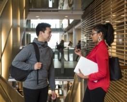 Two students standing inside the FSS building. They are facing each other, the one on the right is talking and the one on the left is smiling attentively. 