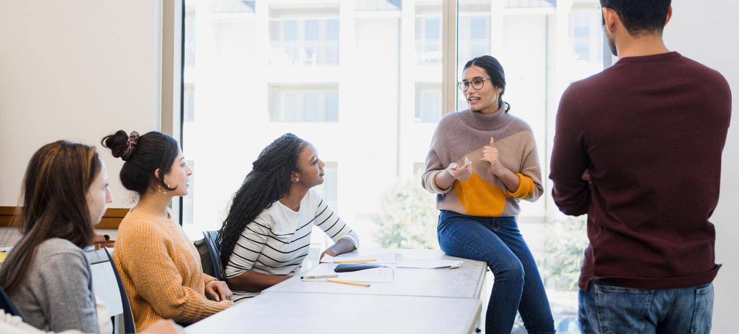 Three criminology students seated and one standing facing another student who is speaking to them in a bright classroom.
