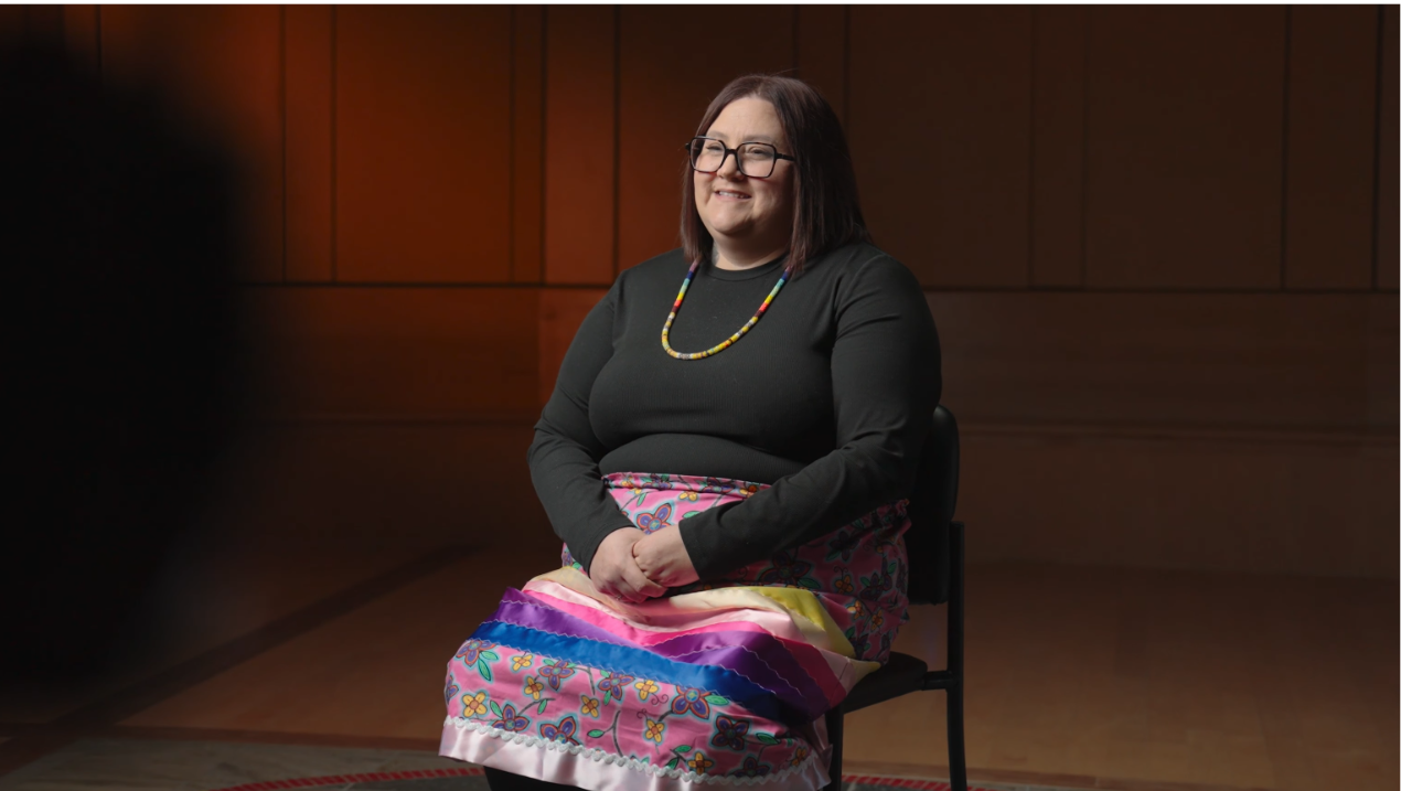 Annie Mailhot wearing traditional ribbon skirt sitting in interview, scene from video about Microprogram.