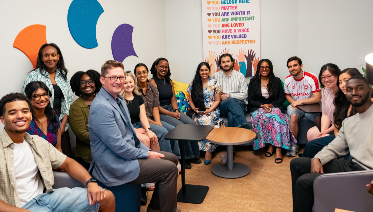 A group of students and faculty members sit together inside the newly opened affinity space, smiling toward the camera. The room features comfortable seating, colourful décor, and an affirmation poster celebrating belonging, respect, and inclusion.