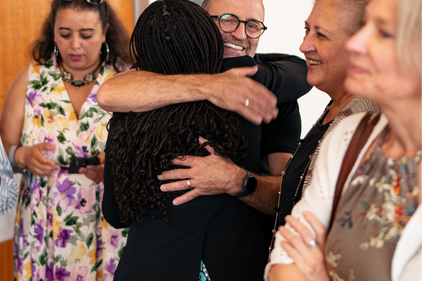 Two attendees share a warm embrace during the opening celebration while other guests gather nearby, chatting and enjoying the event in the university hallway.
