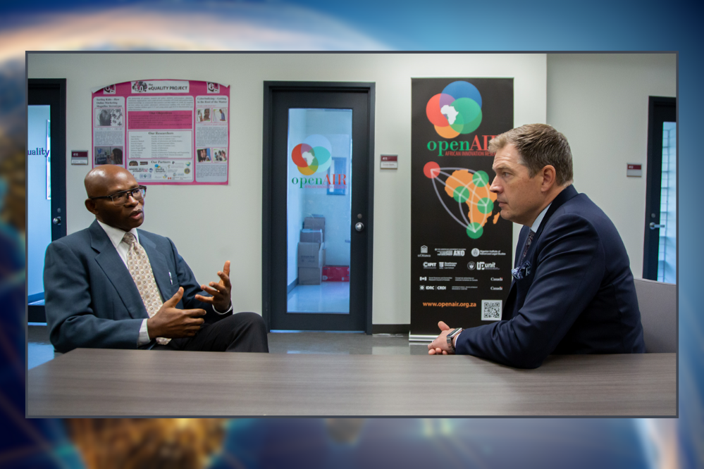 two men sit at a desk, facing one another. They look to be conversing