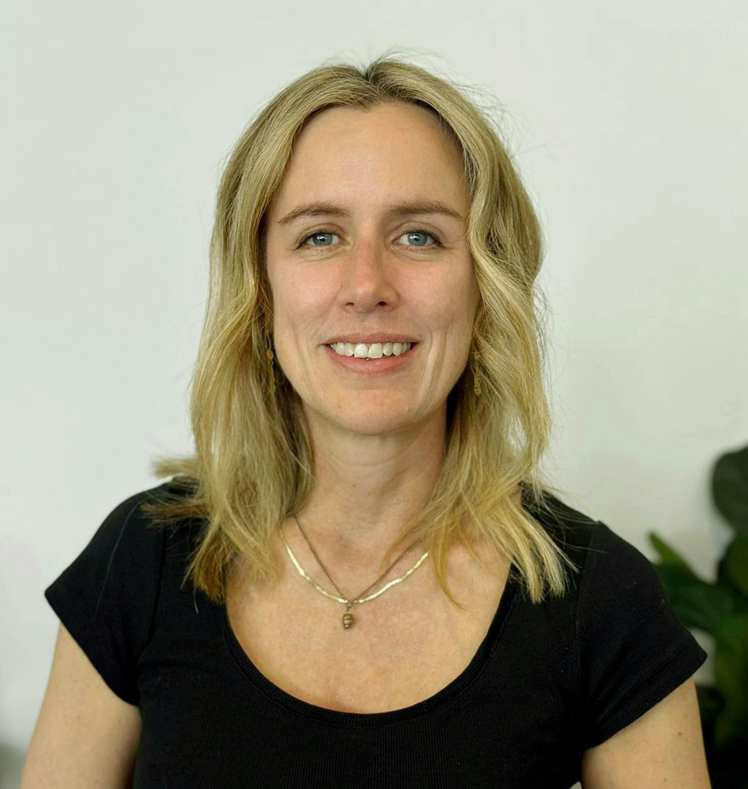  Portrait de la professeure Vanessa Taler souriante, portant un haut noir, avec un arrière-plan uni et une plante partiellement visible.
