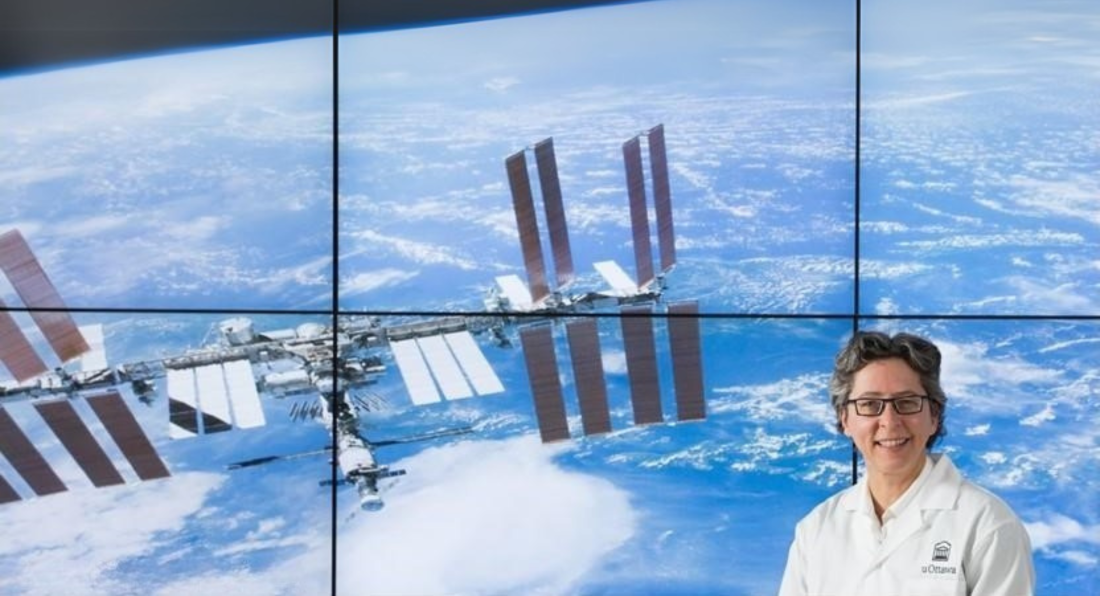 La professeure Odette Laneuville, en sarrau blanc, se tient devant un écran où l’on voit une image de la Station spatiale internationale en orbite autour de la Terre. 